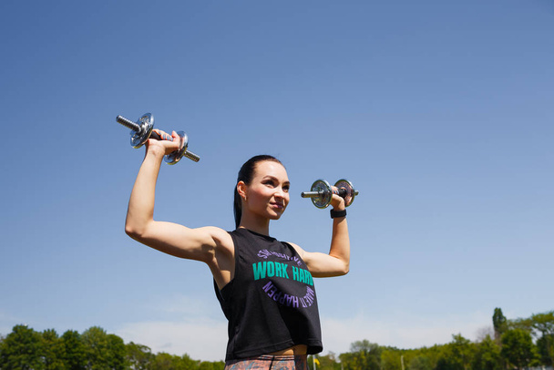 Smiling girl standing on stadium green lawn with dumbbells in hands. Sport, physical training, healthy way of life, fitness concept. - Photo, Image