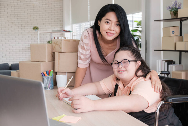 Mère d'âge moyen avec une fille autiste asiatique regarde la caméra et sourit à l'intérieur d'une maison convertie en un petit bureau d'affaires en ligne. Nouvelles affaires à l'ère de l'épidémie COVID-19. - Photo, image