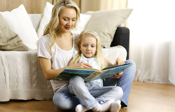 Happy family. Blonde young mother reading a book to her cute daughter while sitting at wooden floor. Motherhood concept - Foto, Imagem