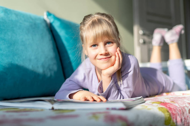 Улыбающийся портрет маленький милый очаровательный счастливый блондинка кавказская девочка нравится лежать на диване кровать и читать сказки книгу. Маленькая девочка читает, мечтает и развивает воображение - Фото, изображение