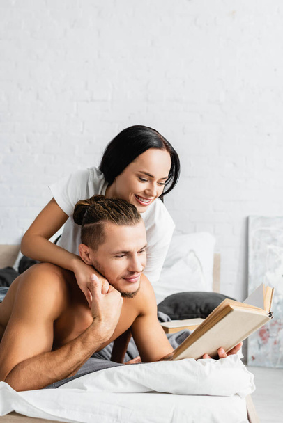 Smiling woman embracing shirtless boyfriend with book on bed  - Foto, imagen