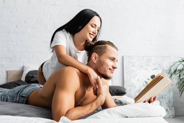 Smiling woman hugging muscular boyfriend reading book on bed  - Фото, зображення
