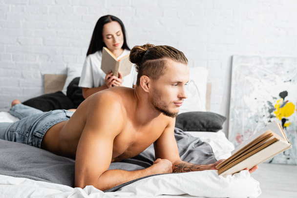 Shirtless man reading book near girlfriend on blurred background in bedroom  - Foto, afbeelding
