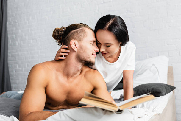 Young woman kissing boyfriend with book on blurred foreground in bedroom  - Фото, изображение