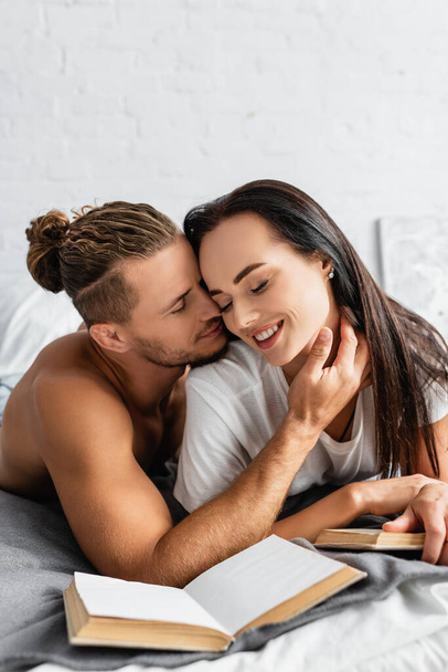 Сексуальный мужчина трогает улыбающуюся женщину возле книг на размытом переднем плане в спальне  - Фото, изображение