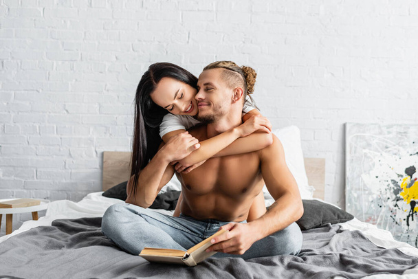 Smiling woman embracing shirtless man holding book on bed  - Photo, Image