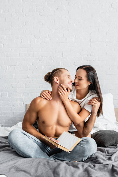 Sexy Mann hält Buch und umarmt fröhliche Freundin auf dem Bett  - Foto, Bild