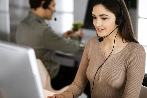 Young friendly girl in headsets is talking to a firms client, while sitting at the desk in a modern office together with her colleague. Call center operators at work - Valokuva, kuva