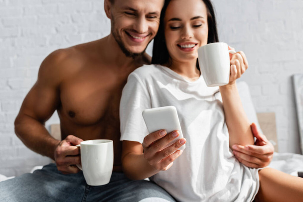 Smiling woman using smartphone near sexy boyfriend with cup on blurred background  - Photo, Image