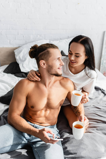 Sexy man holding smartphone near smiling woman with cup on bed  - Photo, Image