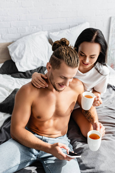 Shirtless man using smartphone near girlfriend with cup on bed  - 写真・画像