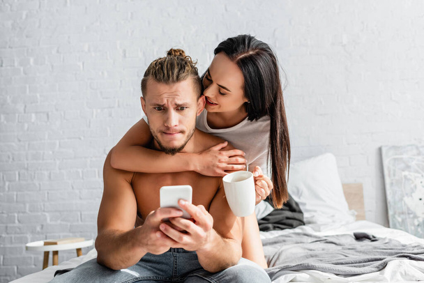 Smiling woman with cup hugging confused boyfriend using smartphone in bedroom  - 写真・画像