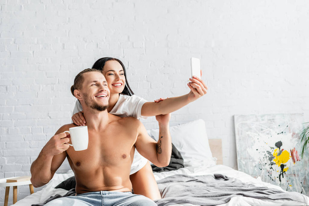 Young woman taking selfie with shirtless boyfriend holding cup on bed  - Photo, Image