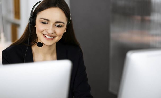 Young friendly girl in headsets is talking to a firms client, while sitting at the desk in a modern office together with her colleague. Call center operators at work - Photo, Image