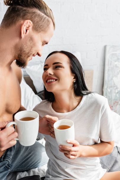 Smiling woman looking at sexy man with cups of tea on bed  - Foto, imagen