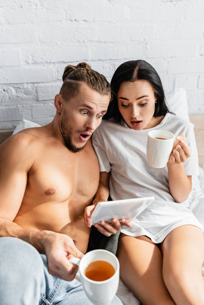 Amazed couple with tea using digital tablet on bed  - Photo, Image