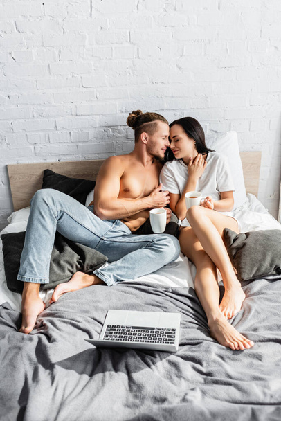 Sexy couple with tea embracing near laptop on bed  - Photo, Image