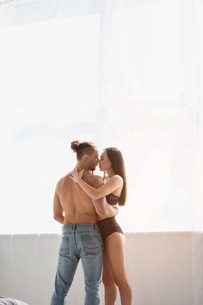 Muscular man in jeans kissing and hugging seductive woman near window at home  - Photo, Image