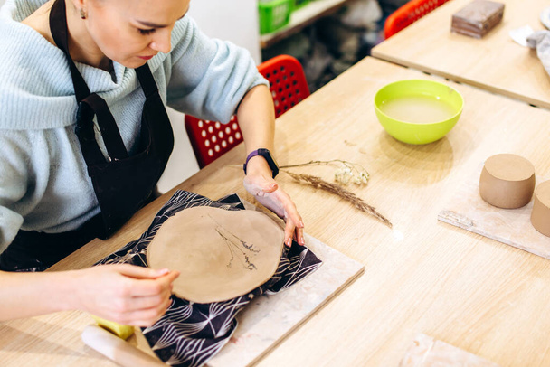 Kobieta przygotowuje glinianą płytę do wypału w pracowni garncarskiej, zdobi talerz z gałązkami lawendy. Widok z góry - Zdjęcie, obraz
