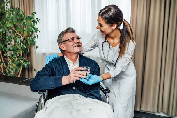 An older man in a wheelchair smiles at the nurse-assistant, she hands him a glass of water. Nursing home, help for the elderly. - Photo, Image