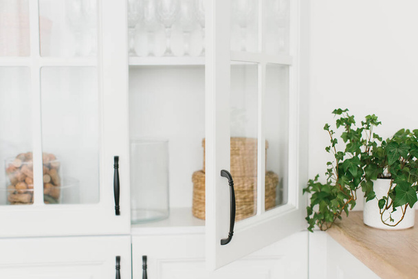 Opened door of white glass cabinet with clean dishes and decor. Scandinavian style kitchen interior. Organization of storage in kitchen. Selective focus. - Photo, image