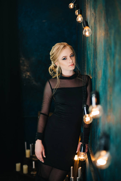 Blonde girl with blue eyes in a black dress in a dark turquoise interior - 写真・画像
