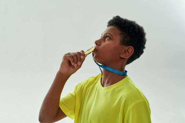 Side view of a teenage african boy kissing gold medal while standing isolated over grey background - Fotografie, Obrázek