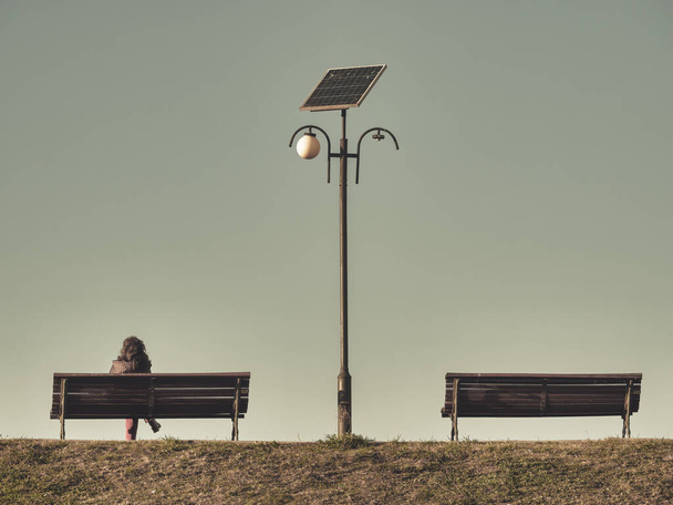 Одинокая женщина сидит на скамейке рядом с фонарным столбом. Две скамейки с фонарным столбом в центре - Фото, изображение