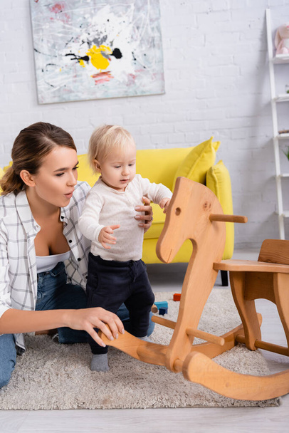 young mother supporting child near rocking horse at home - Foto, Bild