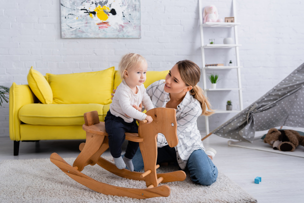 smiling mother supporting sun riding rocking horse at home - Photo, Image