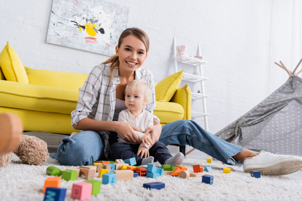 mujer alegre mirando a la cámara mientras juega con cubos de colores en la alfombra con hijo - Foto, imagen