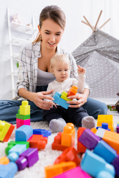 молодая улыбающаяся женщина играет с красочными кубиками конструкции вместе с сыном возле детей wigwam - Фото, изображение