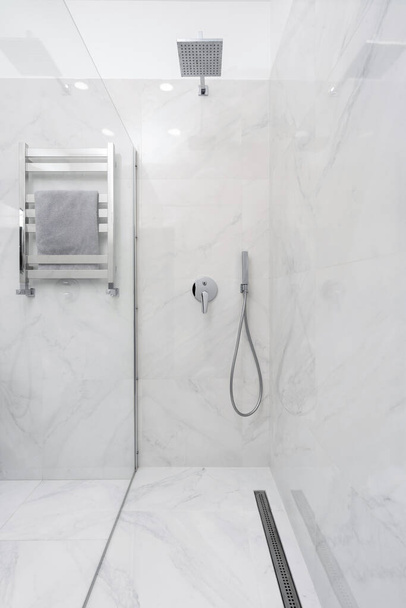 Verticaal uitzicht op moderne badkamer interieur met glazen wand in douchecabine, handdoek op verwarmde rail en sanitair elementen op witte keramische betegelde muur - Foto, afbeelding
