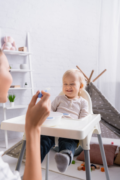 mujer sosteniendo cuchara cerca alegre bebé niño sentado en niños silla, borrosa primer plano - Foto, Imagen
