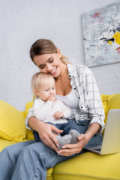 szczęśliwy freelancer obejmujący małego chłopca w pobliżu laptopa na żółtej kanapie - Zdjęcie, obraz