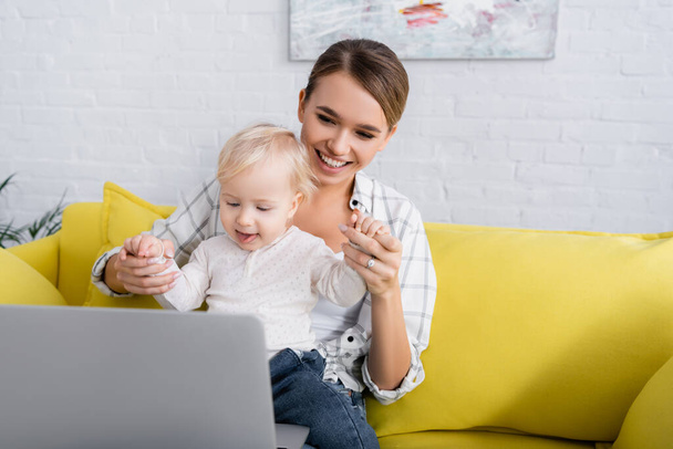 happy freelancer holding hands of little son near laptop on yellow sofa - Photo, Image