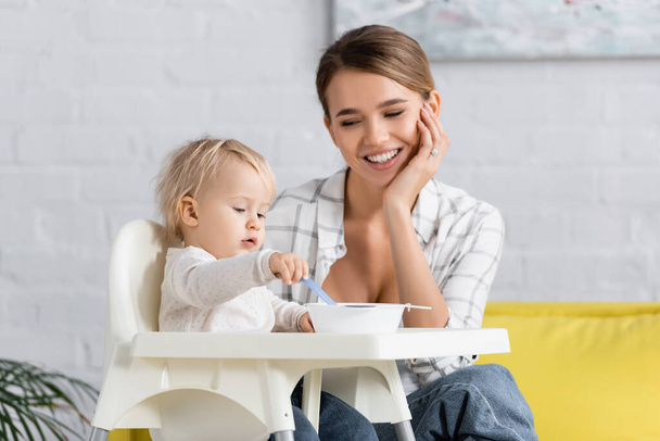 веселая женщина смотрит на маленького сына, держащего ложку, сидя на детском стуле возле чаши - Фото, изображение