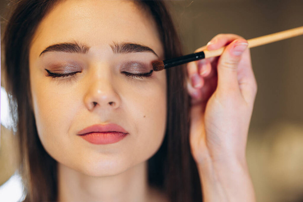 Portrait of a beautiful face of a woman with closed eyes and beautiful makeup. The makeup artist applies the eyeshadow to the eyes with a brush. shining eyeshadow - Foto, Imagem