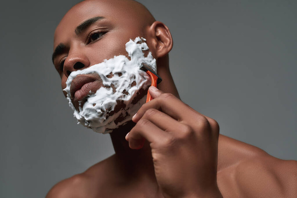 Close up portrait of handsome young african american man with foam applied on his face using steel razor for shaving isolated over gray background - Fotografie, Obrázek