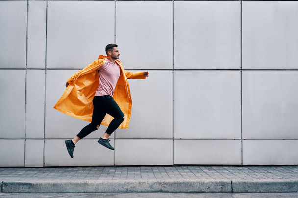 Trendy young man in yellow raincoat jumping in the air on wall street urban background - Foto, Bild
