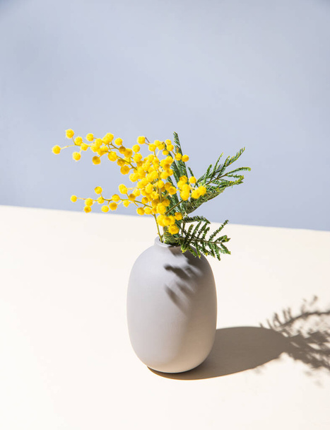 un ramo de flores de mimosa amarilla se encuentra en un jarrón de cerámica con sombra sobre un fondo amarillo y gris. concepto del 8 de marzo, feliz día de la mujer. espacio de copia - Foto, Imagen