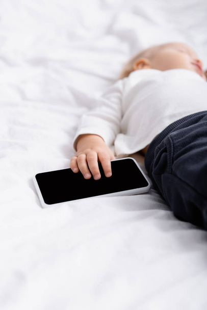 背景にぼやけた状態で寝ている子供の近くに空白の画面を持つスマートフォンの - 写真・画像