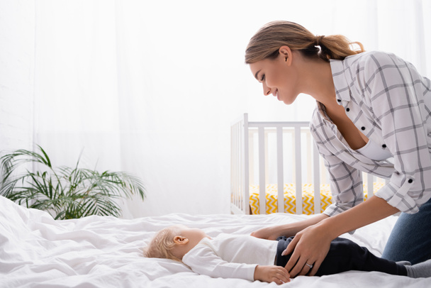 Yatak odasında küçük oğlunun üzerine eğilmiş genç bir kadının yan görünüşü - Fotoğraf, Görsel
