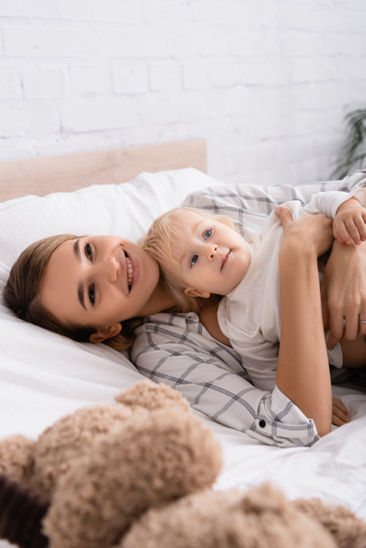 happy woman embracing little son while lying in bedroom on blurred foreground - Фото, изображение