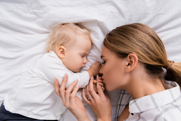 Ansicht der jungen Frau, die die Hand ihres Sohnes küsst, der auf weißem Bettzeug schläft - Foto, Bild