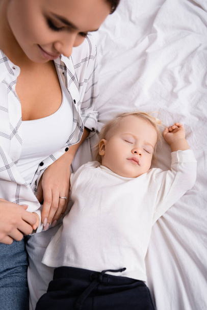 над головой вид улыбающейся женщины касающейся руки спящего сына, размытый передний план - Фото, изображение