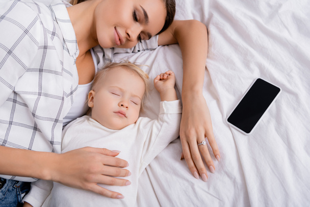 vista aérea de la mujer sonriente acostada cerca del bebé y el teléfono inteligente con pantalla en blanco - Foto, imagen