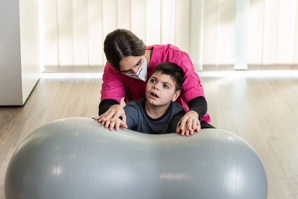 mozgássérült gyermek és fizioterapeuta egy mogyoró tornatermi bálon egyensúlyozó gyakorlatokat végez. pandémiás maszkvédelem - Fotó, kép