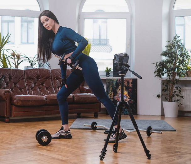 Mujer de fitness sosteniendo una mancuerna posa en la cámara en el apartamento - Foto, Imagen
