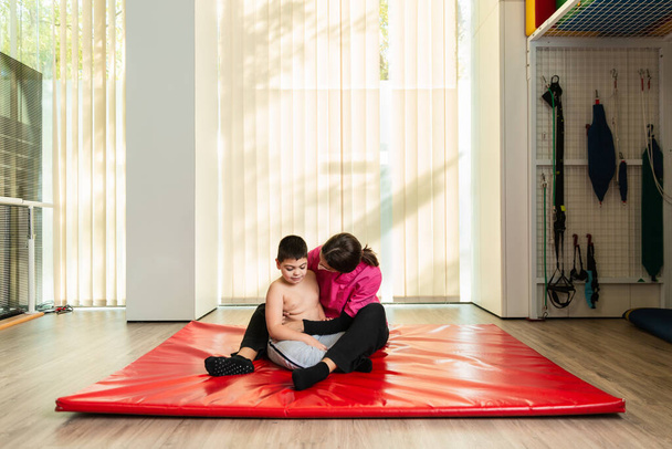 behindertes Kind und Physiotherapeut auf einer roten Gymnastikmatte bei Übungen. Pandemieschutz - Foto, Bild